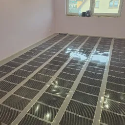 Rozłożona folia grzewcza na podłodze w pomieszczeniu z różowymi ścianami, przygotowana do dalszych prac wykończeniowych, widok na okno z zewnątrz.