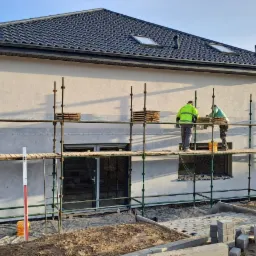 Dwóch pracowników na rusztowaniu podczas ocieplania elewacji domu jednorodzinnego, widoczne płyty styropianowe i puszki z klejem