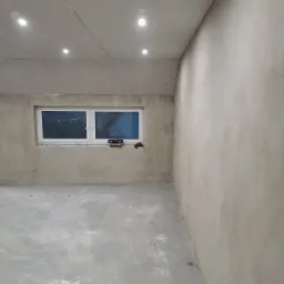 Surowe wnętrze pokoju w trakcie prac wykończeniowych: ściany pokryte gładzią, sufit z płyt kartonowo-gipsowych z zamontowanym oświetleniem punktowym, okno z narzędziami na parapecie.