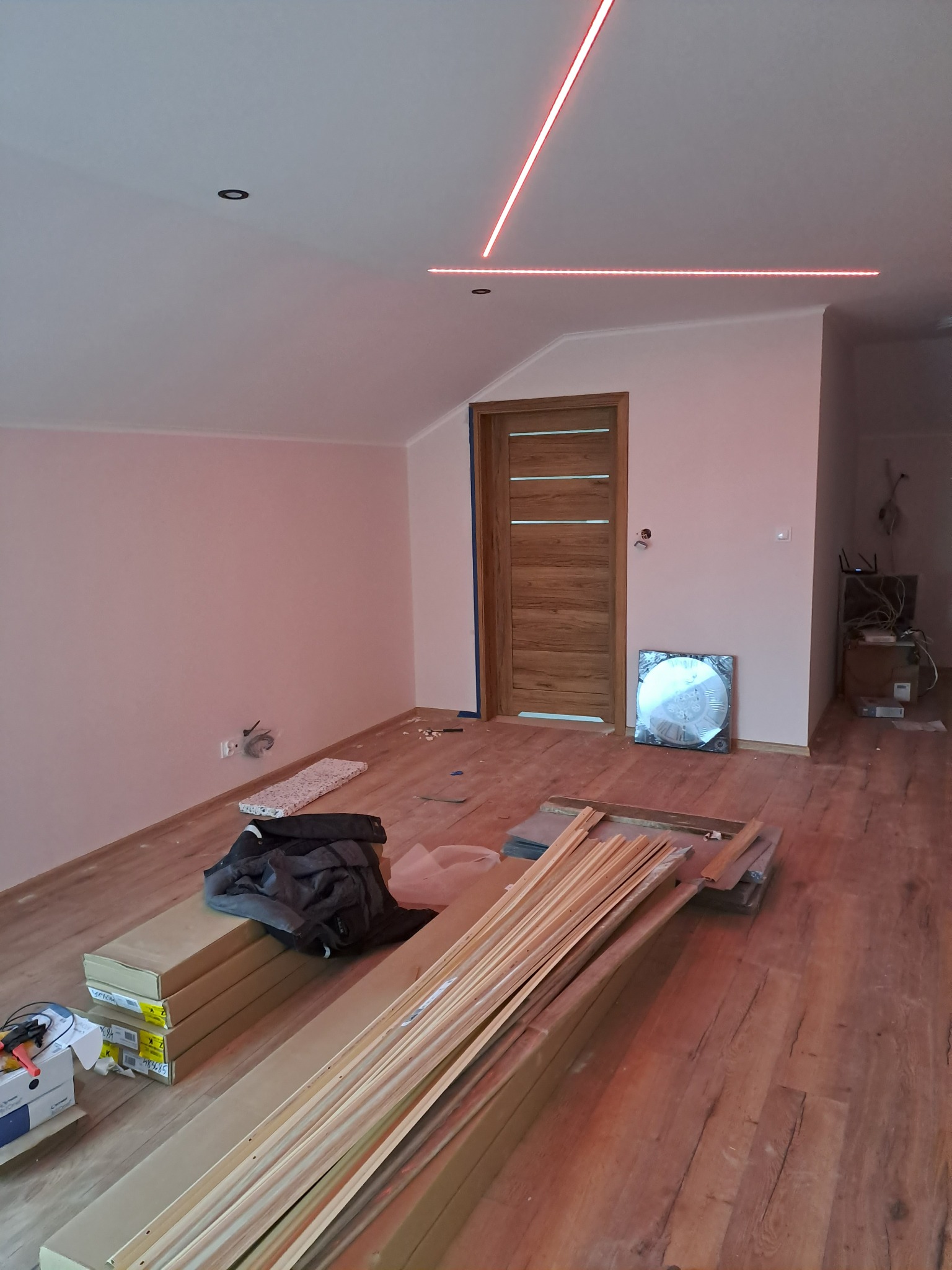 Pokój na poddaszu w trakcie prac wykończeniowych, widoczne profile do płyt kartonowo-gipsowych, oświetlenie LED na suficie i drewniana podłoga.