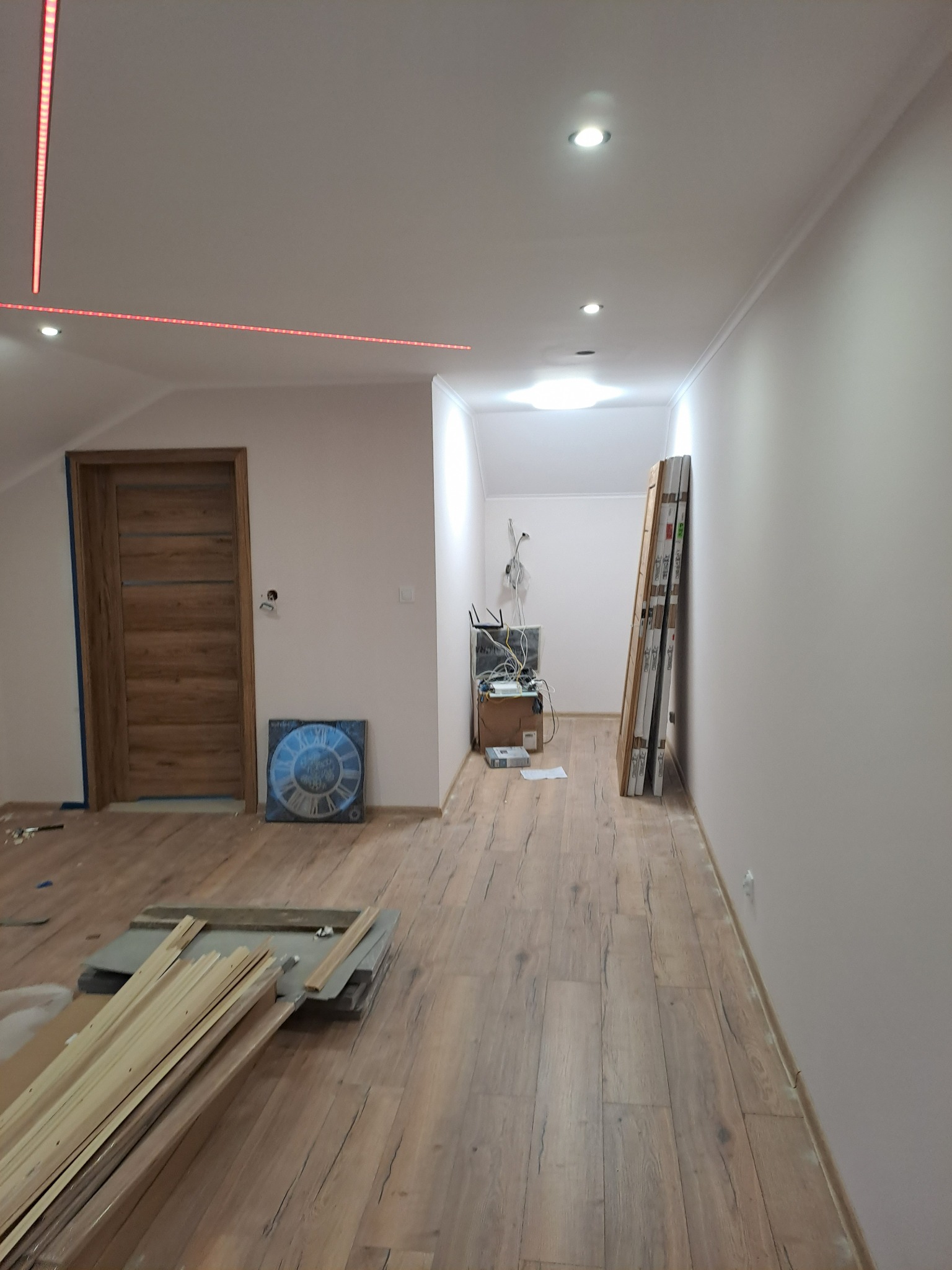 Wnętrze pokoju w trakcie prac wykończeniowych: położona podłoga, zamontowane drzwi, widoczne listwy LED na suficie i materiały budowlane na podłodze.