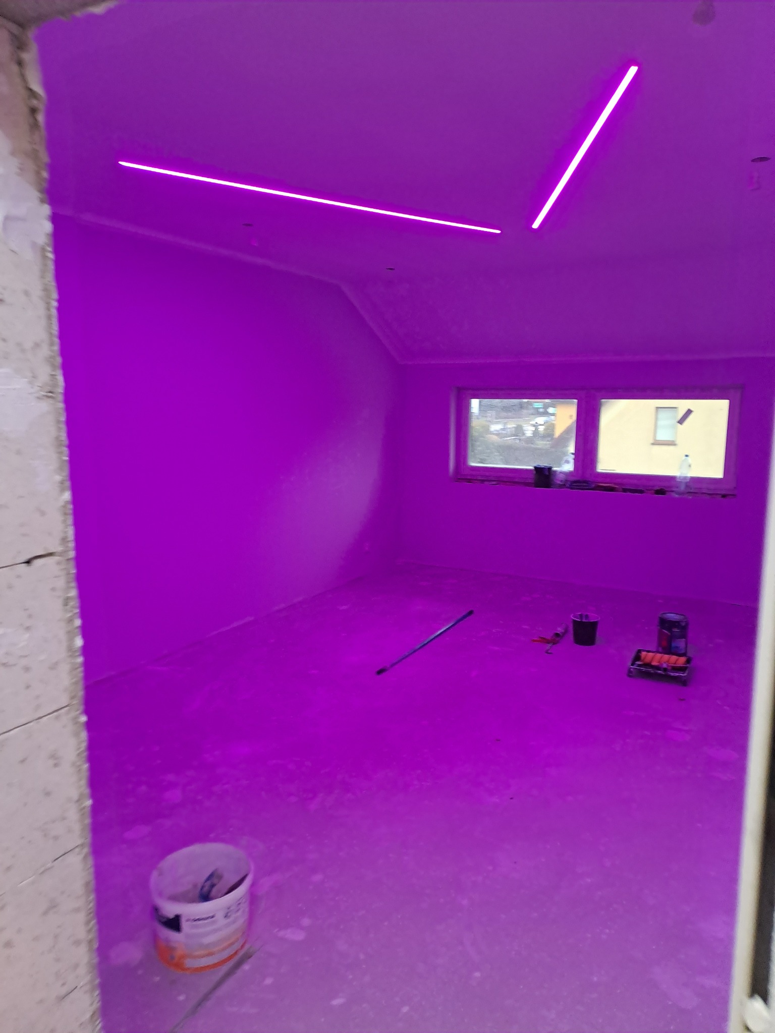 Pokój po gładzi, oświetlony fioletowym światłem LED, z widocznymi narzędziami malarskimi i wiadrem z farbą, przygotowany do dalszych prac wykończeniowych.