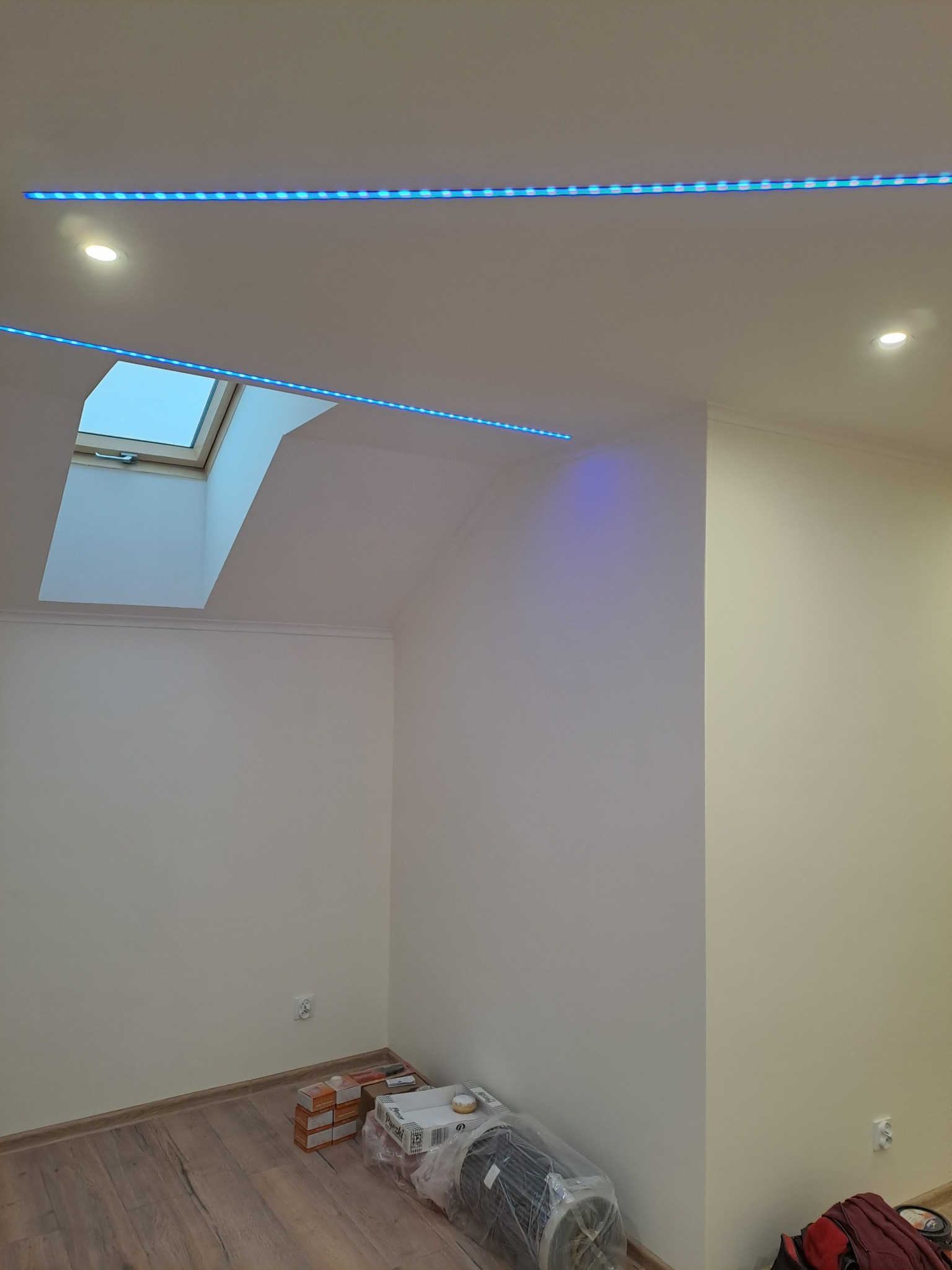 Pokój na poddaszu z oknem dachowym i oświetleniem LED, widoczne materiały budowlane i narzędzia na podłodze.