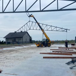 KORDALSKI - Żuraw teleskopowy montuje stalową konstrukcję dachu hali na betonowym fundamencie. W tle dom jednorodzinny i pochmurne niebo.