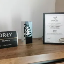Nagrody i dyplom Kancelarii Radcy Prawnego Monika Przyszlak, w tym statuetka Orły Prawa i dyplom za zajęcie II miejsca w Człuchowie w 2021 roku, ustawione na drewnianej komodzie.