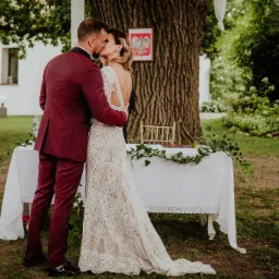 Para młoda całuje się podczas ceremonii ślubnej na świeżym powietrzu, na tle drzewa i stołu z białym obrusem udekorowanym bluszczem, w tle budynek i godło Polski.