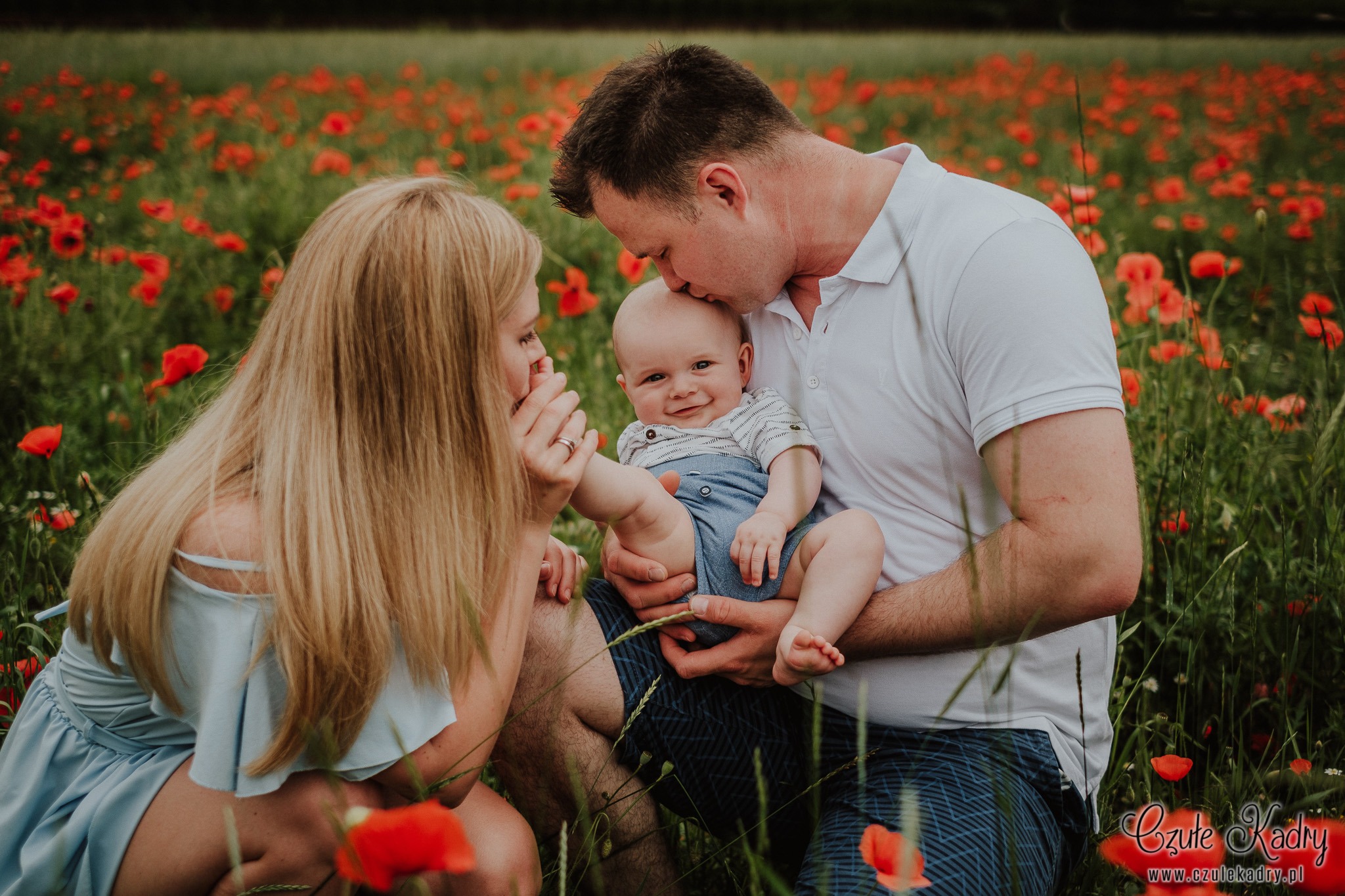 Rodzinne ujęcie w plenerze: mama w błękitnej sukience trzyma dłoń na stopie uśmiechniętego niemowlęcia, tata całuje dziecko w czoło na tle kwitnącego pola maków.