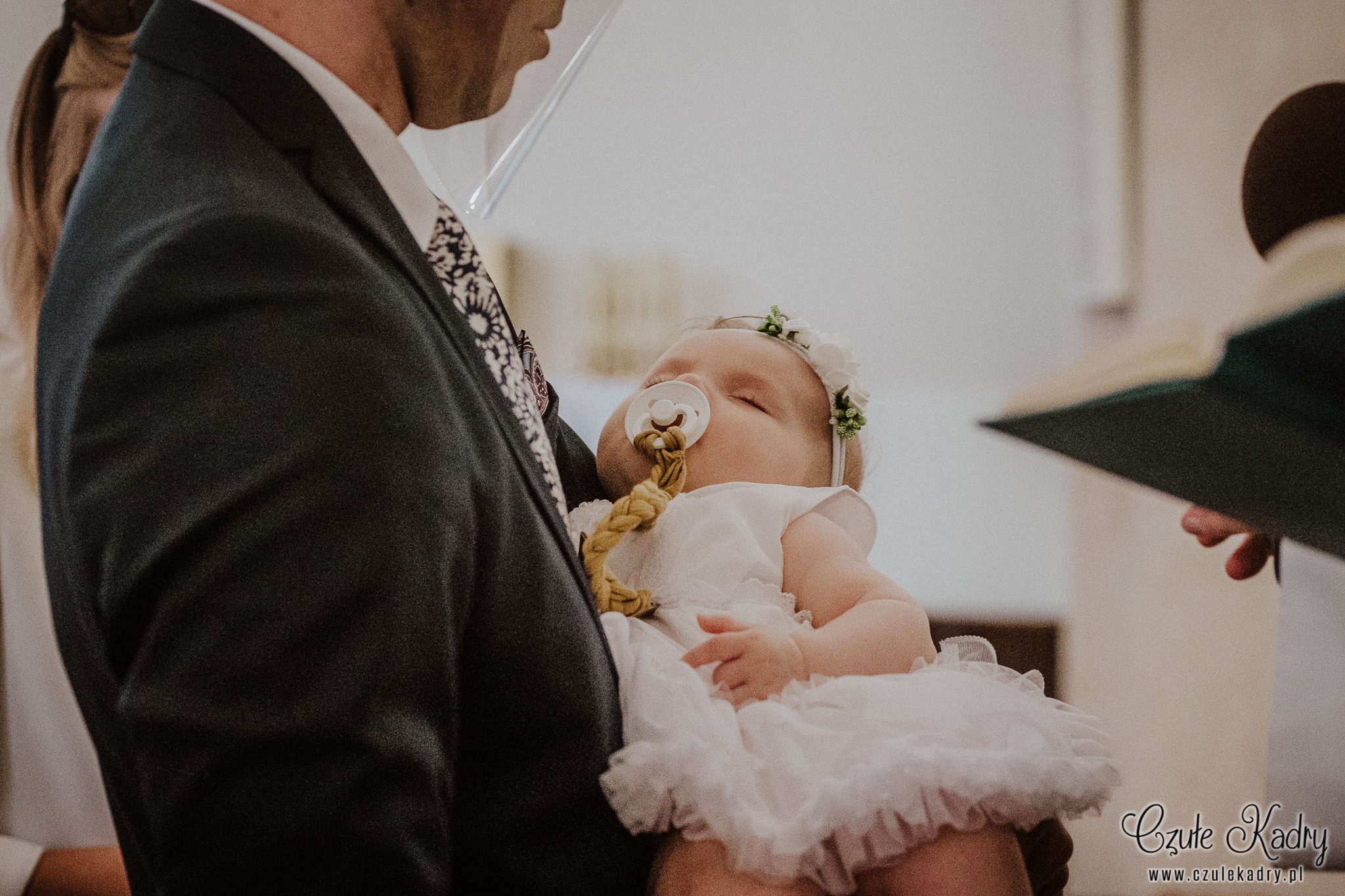 Śpiące niemowlę w białej sukience i wianku na głowie, trzymane na rękach przez mężczyznę w garniturze podczas ceremonii, z widocznym fragmentem księgi i dłonią w tle.