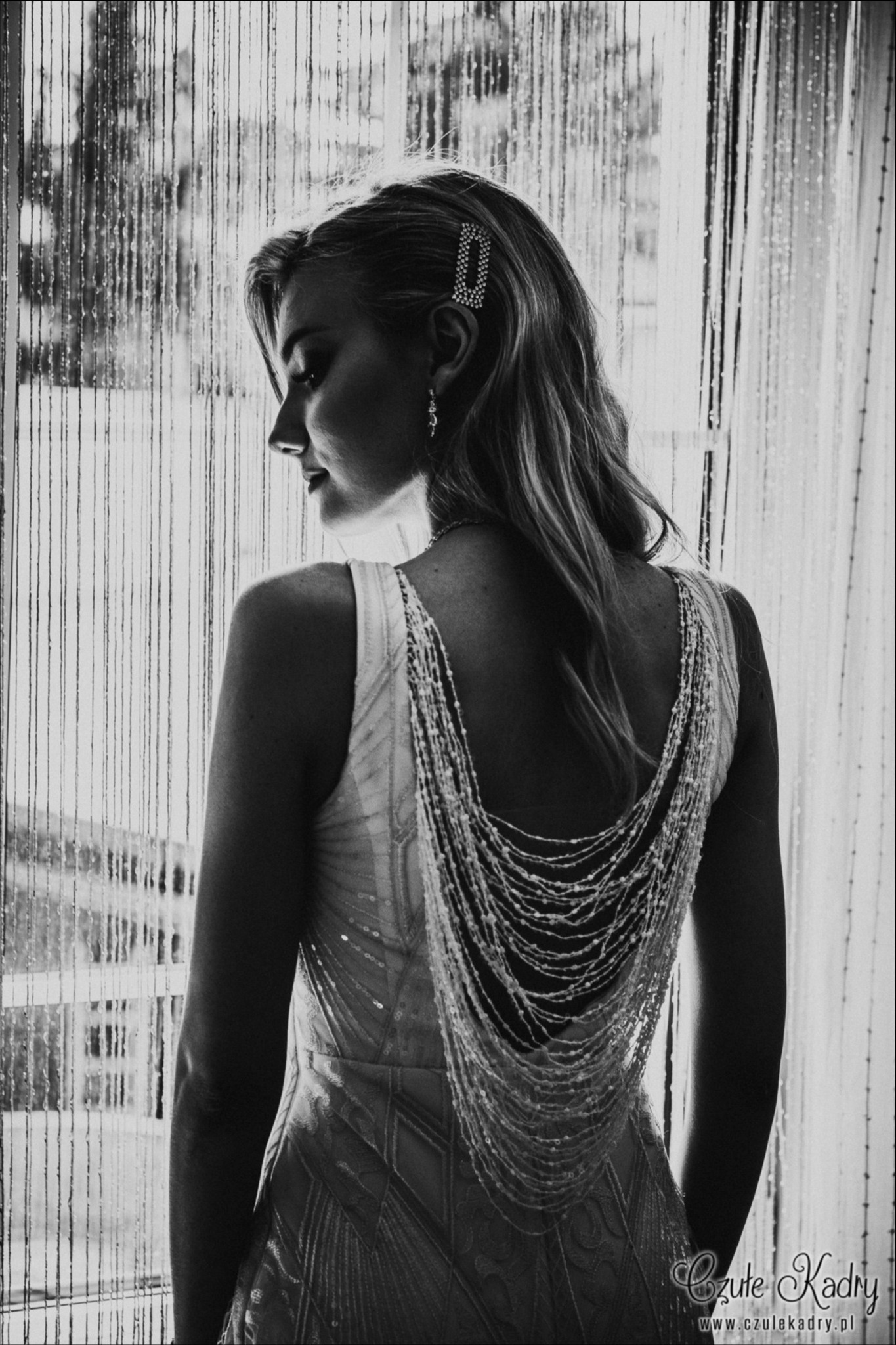 Elegancka kobieta w sukni z kaskadą pereł na plecach, z ozdobną spinką we włosach, patrzy w bok na tle okna z deszczową zasłoną, czarno-białe ujęcie.