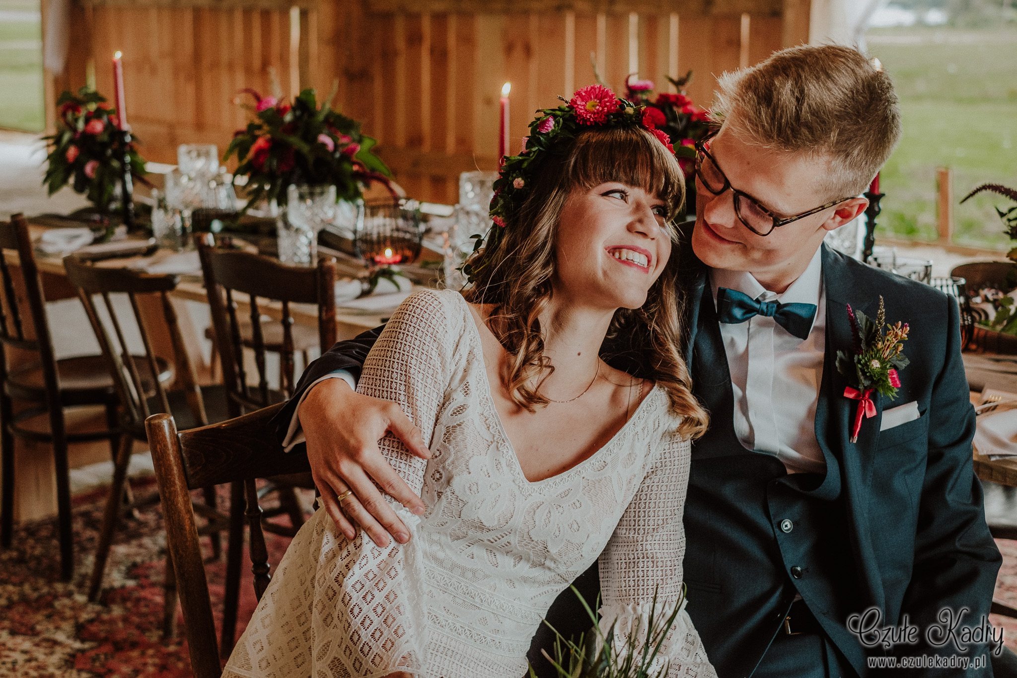 Uśmiechnięta panna młoda w wianku z kwiatów, spoglądająca na pana młodego w okularach i ciemnym garniturze z muszką, siedzą obok stołu z dekoracjami kwiatowymi i świecami, w rustykalnym wnętrzu.