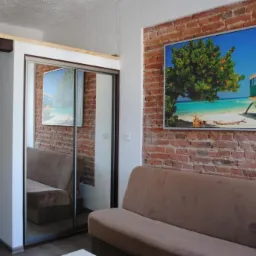 Wnętrze pokoju z sofą na tle ściany z czerwonej cegły, na której wisi obraz z motywem plaży; obok szafa z przesuwnymi drzwiami lustrzanymi.