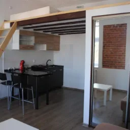 Wnętrze mieszkania z antresolą sypialnianą, dostępną po drewnianej drabinie, z widoczną kuchnią i salonem oddzielonym przesuwnymi drzwiami z lustrem, w którym odbija się fragment ściany z cegły.