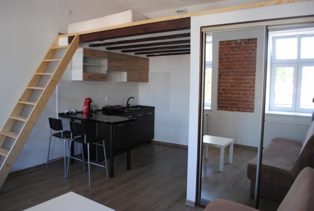 Wnętrze mieszkania z antresolą sypialnianą, dostępną po drewnianej drabinie, z widoczną kuchnią i salonem oddzielonym przesuwnymi drzwiami z lustrem, w którym odbija się fragment ściany z cegły.