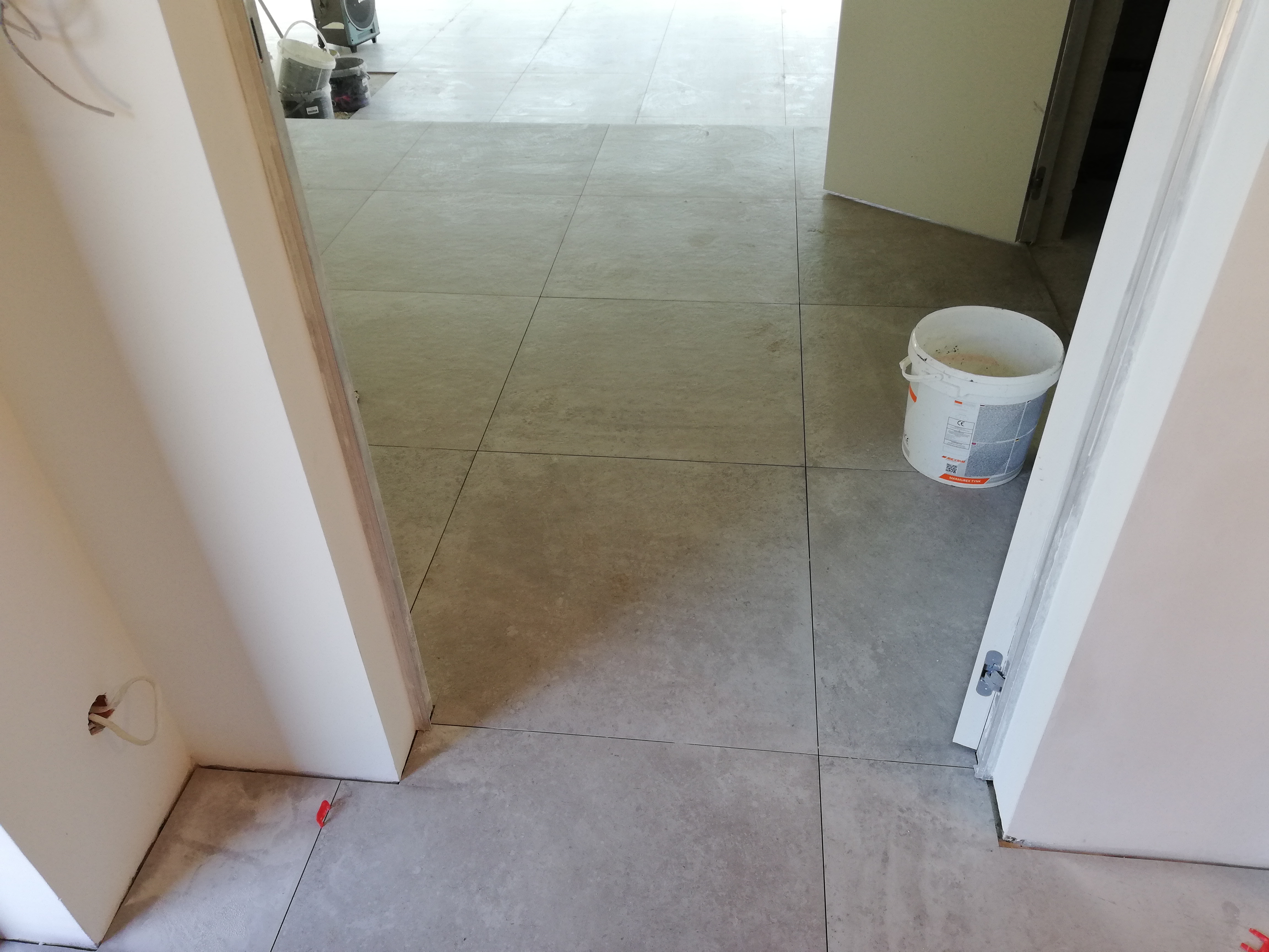 Ułożone szare płytki podłogowe w nowym pomieszczeniu, widoczne białe, świeżo malowane ościeżnice drzwiowe, wiadro z zaprawą i czerwone kliny poziomujące.