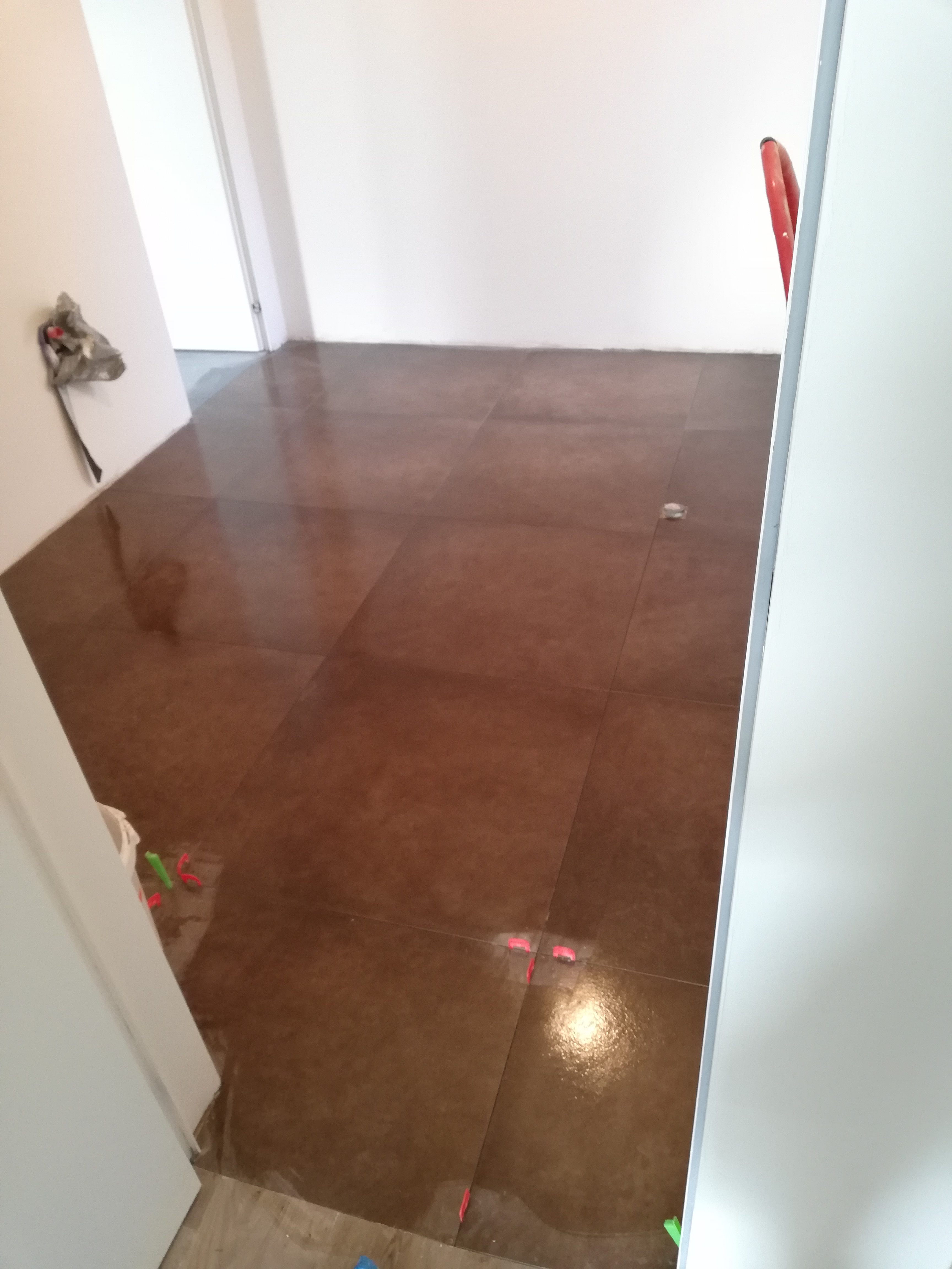 Świeżo ułożone, brązowe płytki podłogowe z plastikowymi krzyżykami dystansowymi w jasnym pomieszczeniu, widok zza białych drzwi.