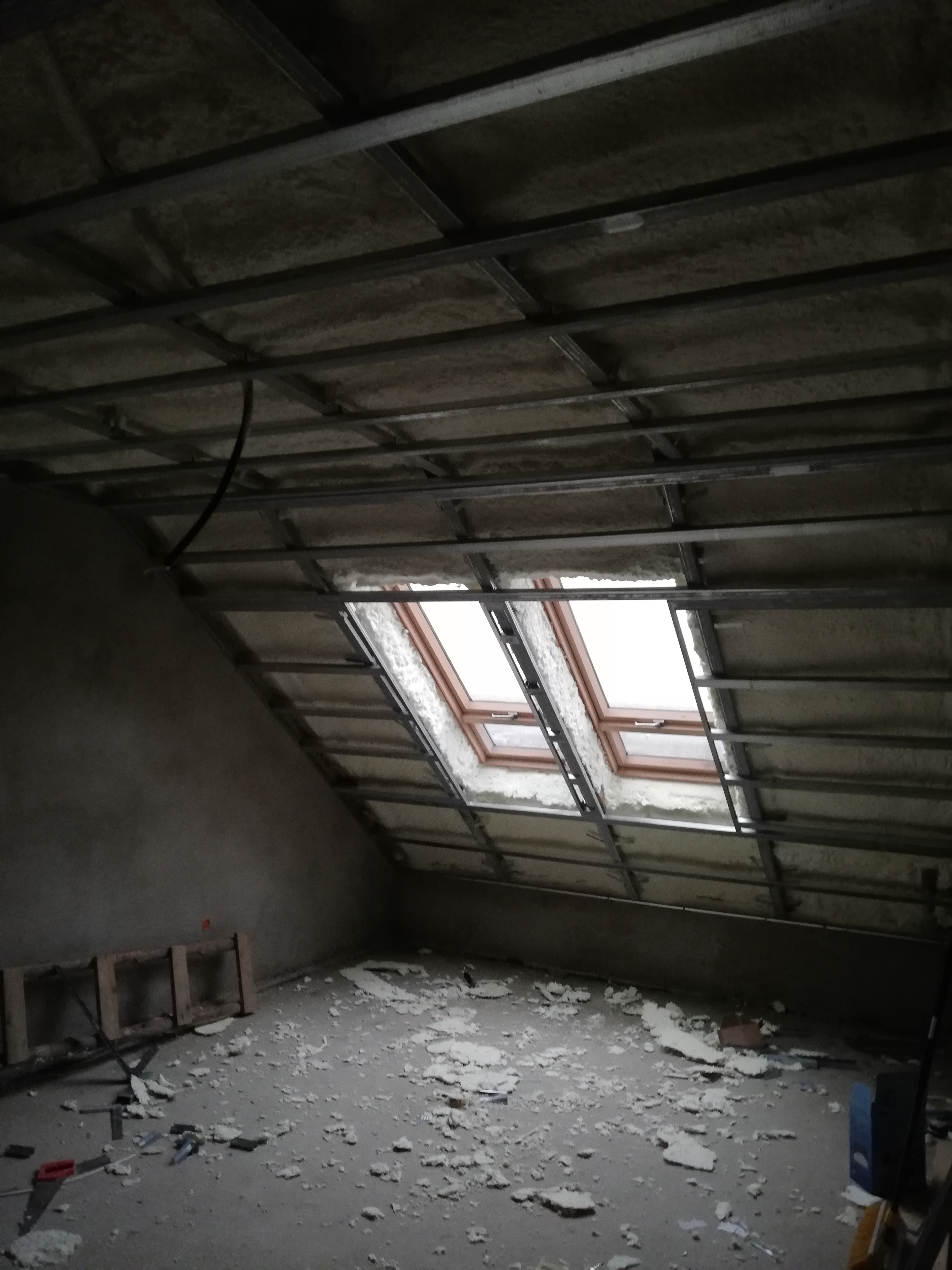 Poddasze w trakcie ocieplania, widoczne dwa okna dachowe, metalowa konstrukcja sufitu podwieszanego oraz resztki materiału izolacyjnego na podłodze.