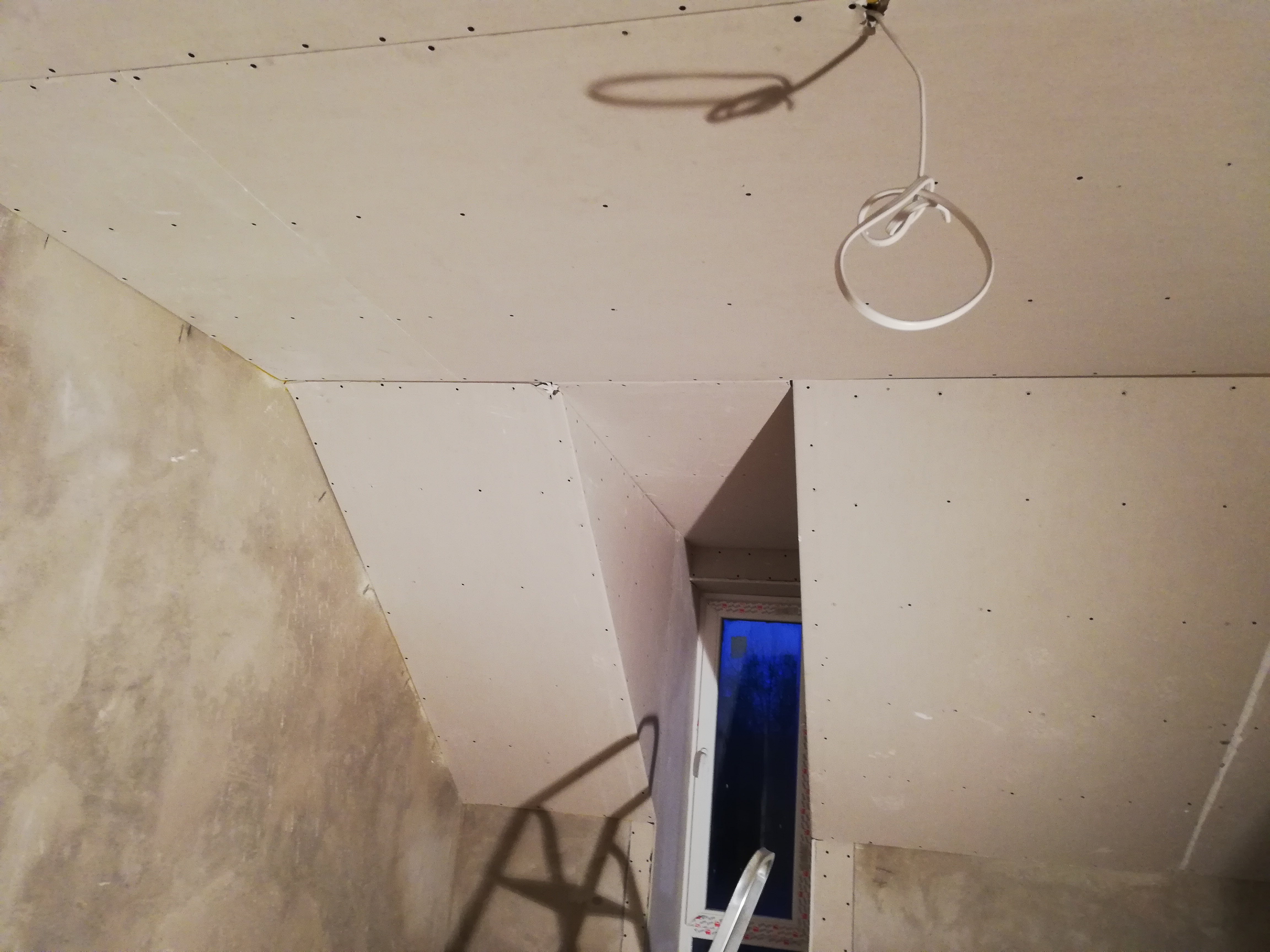 Ściany i sufit w trakcie montażu płyt gipsowo-kartonowych, widoczne łączenia płyt i wkręty, fragment okna i przewód elektryczny zwisający z sufitu.