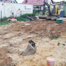 Żółta koparka Volvo na placu budowy, świeżo wykopana ziemia z widocznymi śladami opon, w tle biały kontener z logo firmy i zielony kontener magazynowy.