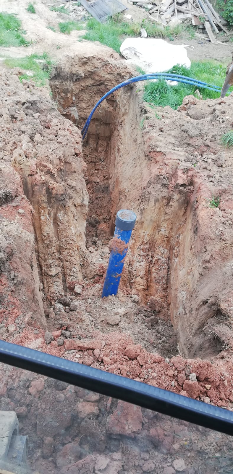 Wykopany wąski rów w ziemi z widocznymi ścianami z gliny i piasku, wewnątrz pionowo umieszczona niebieska rura z szarym deklem, u góry widoczne niebieskie rury doprowadzające wodę.