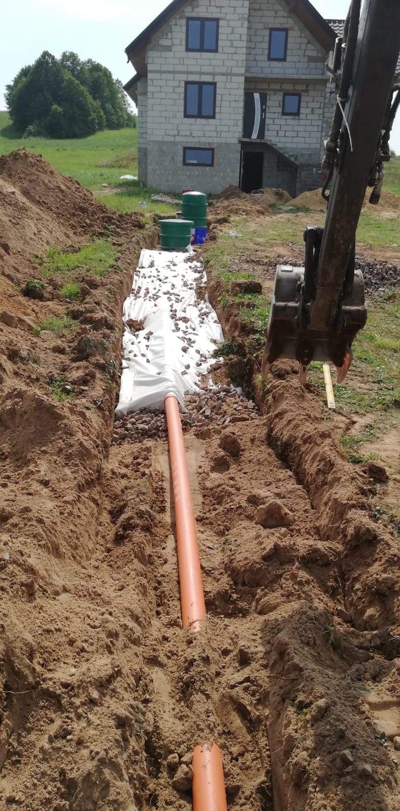 Wykopany rów z pomarańczową rurą drenażową ułożoną na białej geowłókninie z warstwą kamieni, w tle koparka i budynek mieszkalny z zielonymi zbiornikami.