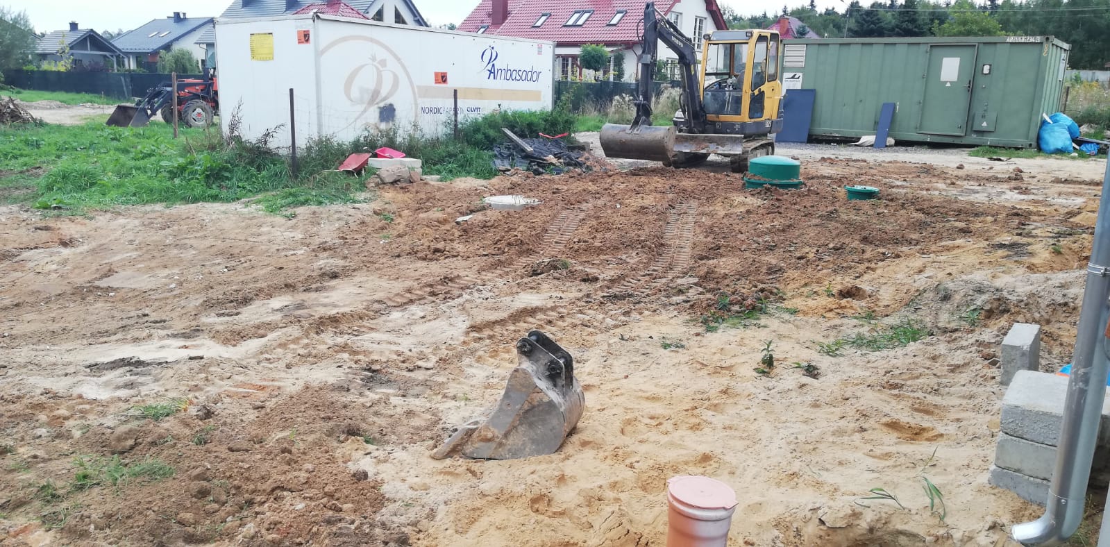 Żółta koparka Volvo na placu budowy, świeżo wykopana ziemia z widocznymi śladami opon, w tle biały kontener z logo firmy i zielony kontener magazynowy.