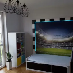 Pokój dziecięcy z łóżkiem i fototapetą przedstawiającą stadion, obramowaną czarno-białymi pasami i niebieskim oświetleniem LED.