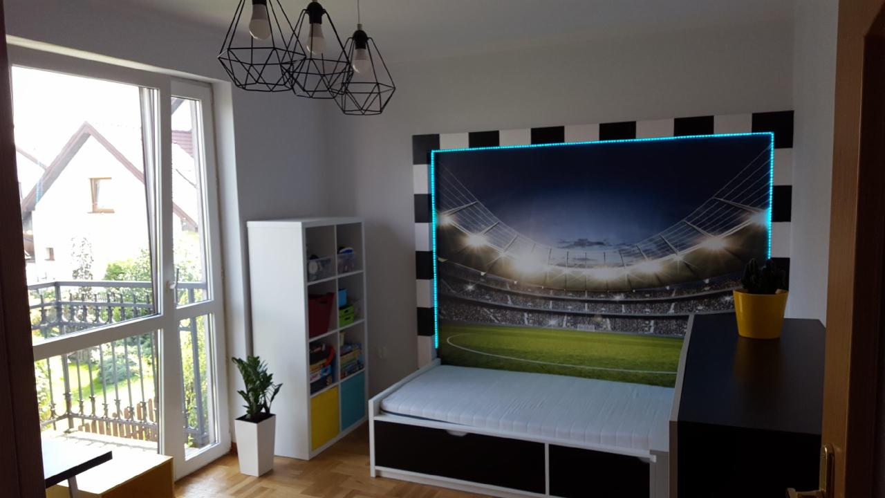 Pokój dziecięcy z łóżkiem i fototapetą przedstawiającą stadion, obramowaną czarno-białymi pasami i niebieskim oświetleniem LED.