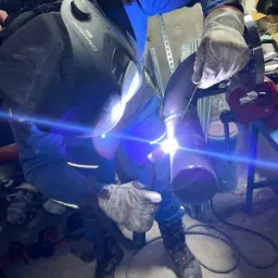 Spawacz w masce 3M Speedglas D100 EX, w roboczych rękawicach, spawa rurę w warsztacie, widoczny intensywny błysk spawania.