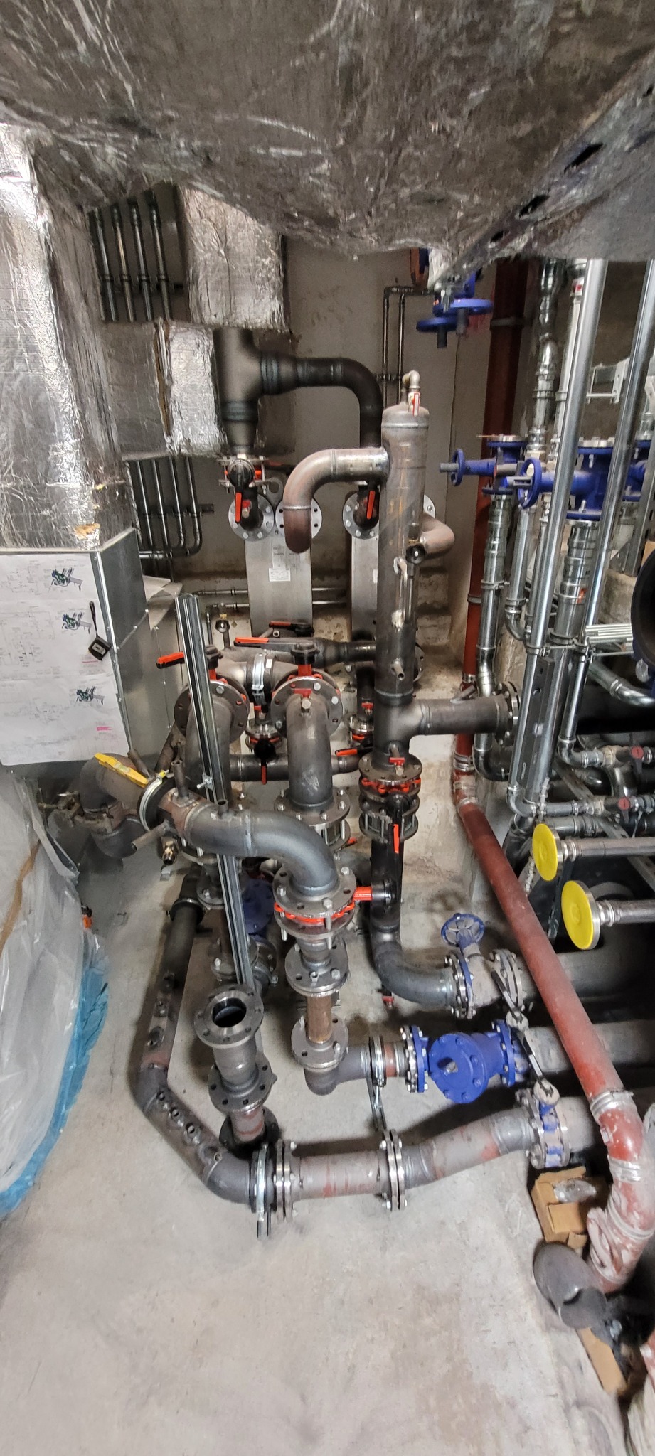 Złożona instalacja rurowa z licznymi zaworami i złączkami w pomieszczeniu technicznym z izolacją na suficie, widoczne elementy spawane i ślady po spawaniu.