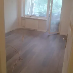 Wnętrze pokoju po remoncie z nową szarą podłogą z paneli, świeżo pomalowanymi ścianami i otwartym balkonem z widokiem na zieleń za oknem.