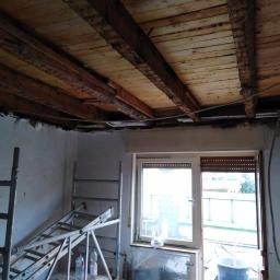 Grzegorz Serdeń - Wnętrze w trakcie demontażu sufitu, widoczne odsłonięte drewniane belki stropowe, drabiny i butelki z wodą w pomieszczeniu z oknem.