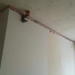 Laserowy niwelator przymocowany do ściany z widoczną czerwoną linią lasera, surowy strop z widocznymi śladami po zalaniach, przygotowania do prac wykończeniowych.