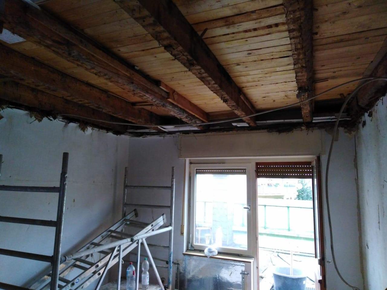 Wnętrze w trakcie demontażu sufitu, widoczne odsłonięte drewniane belki stropowe, drabiny i butelki z wodą w pomieszczeniu z oknem.