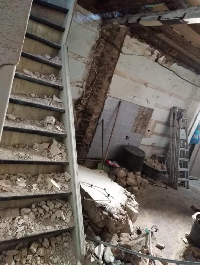 Wnętrze w trakcie demontażu: schody zasypane gruzem, odsłonięta cegła, fragmenty płytek na ścianie pod skosem dachu, drabina oparta o ścianę, widoczne elementy instalacji elektrycznej.