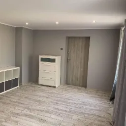 Wnętrze pokoju po malowaniu ścian na szaro, widoczne panele podłogowe imitujące drewno, białe meble, drzwi i zasłony.