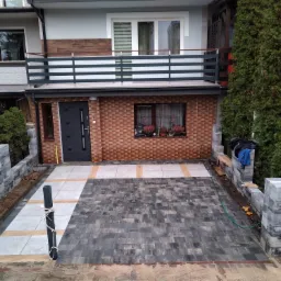 Ułożona, szara kostka na podjeździe domu jednorodzinnego z elementami jasnych płyt, widoczne obrzeża i częściowo zagospodarowany teren wokół.