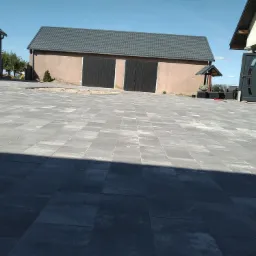 Szara nawierzchnia z prostokątnych płyt ułożona przed budynkami gospodarczymi z ciemnymi bramami, widok na posesję w słoneczny dzień.