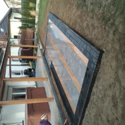 Nowa nawierzchnia z szarych płyt i ciemnych obrzeży na przygotowanym podłożu obok domu z drewnianą konstrukcją tarasu.