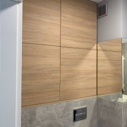 Euro Expo - Drewniane szafki w zabudowie nad toaletą w łazience, z widocznym przyciskiem spłuczki i fragmentem lustra.