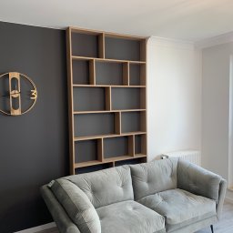 Euro Expo - Nowoczesny salon w Poznaniu z szarą sofą i drewnianym regałem na książki o asymetrycznych półkach, na ścianie zegar w stylu loft.