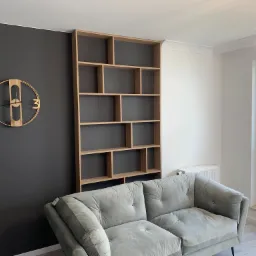 Nowoczesny salon w Poznaniu z szarą sofą i drewnianym regałem na książki o asymetrycznych półkach, na ścianie zegar w stylu loft.