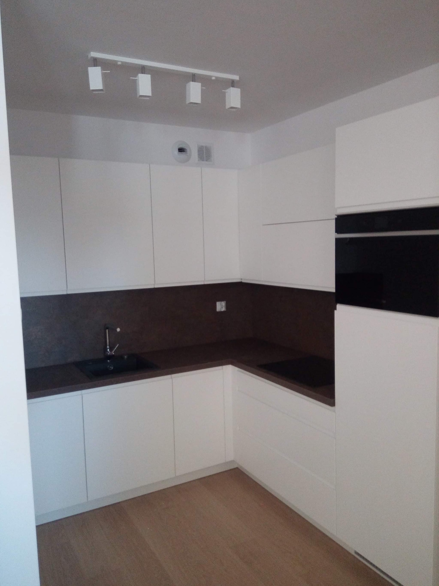 Minimalistyczna, biała zabudowa kuchenna z ciemnym blatem i oświetleniem punktowym na suficie.