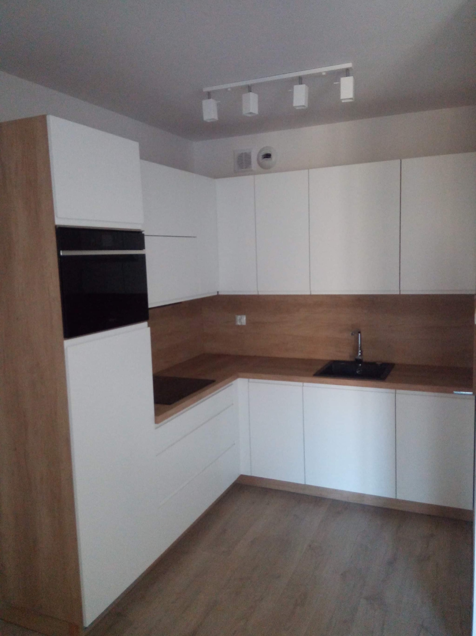 Nowoczesna, jasna kuchnia z białymi frontami szafek i drewnianym blatem oraz zabudowaną czarną płytą grzewczą i piekarnikiem, oświetlona listwą z reflektorami.