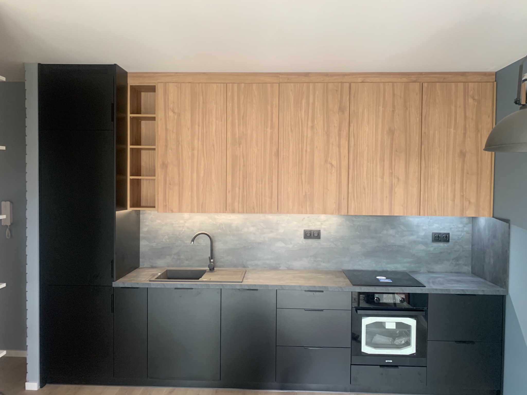 Nowoczesna kuchnia z czarnymi szafkami dolnymi i drewnianymi górnymi, szary blat i backsplash, minimalistyczny design, oświetlenie LED pod szafkami górnymi.