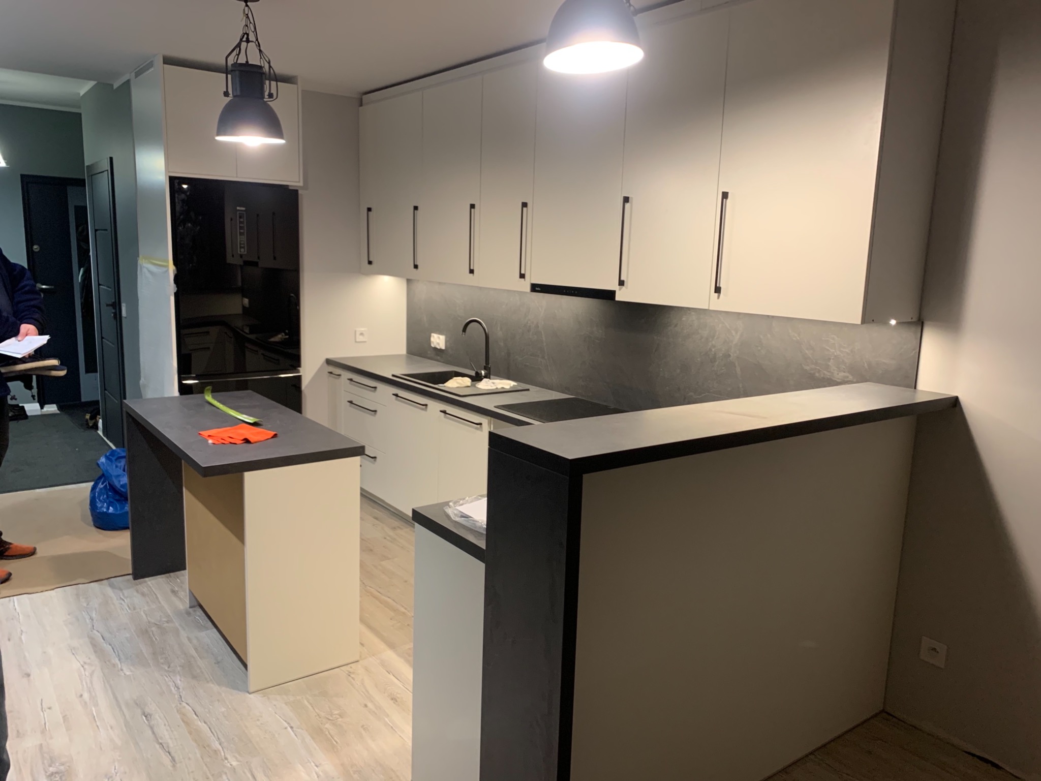 Nowoczesna kuchnia z białymi szafkami, czarnymi uchwytami, szarym blatem i wyspą kuchenną z pomarańczowymi akcentami na blacie.