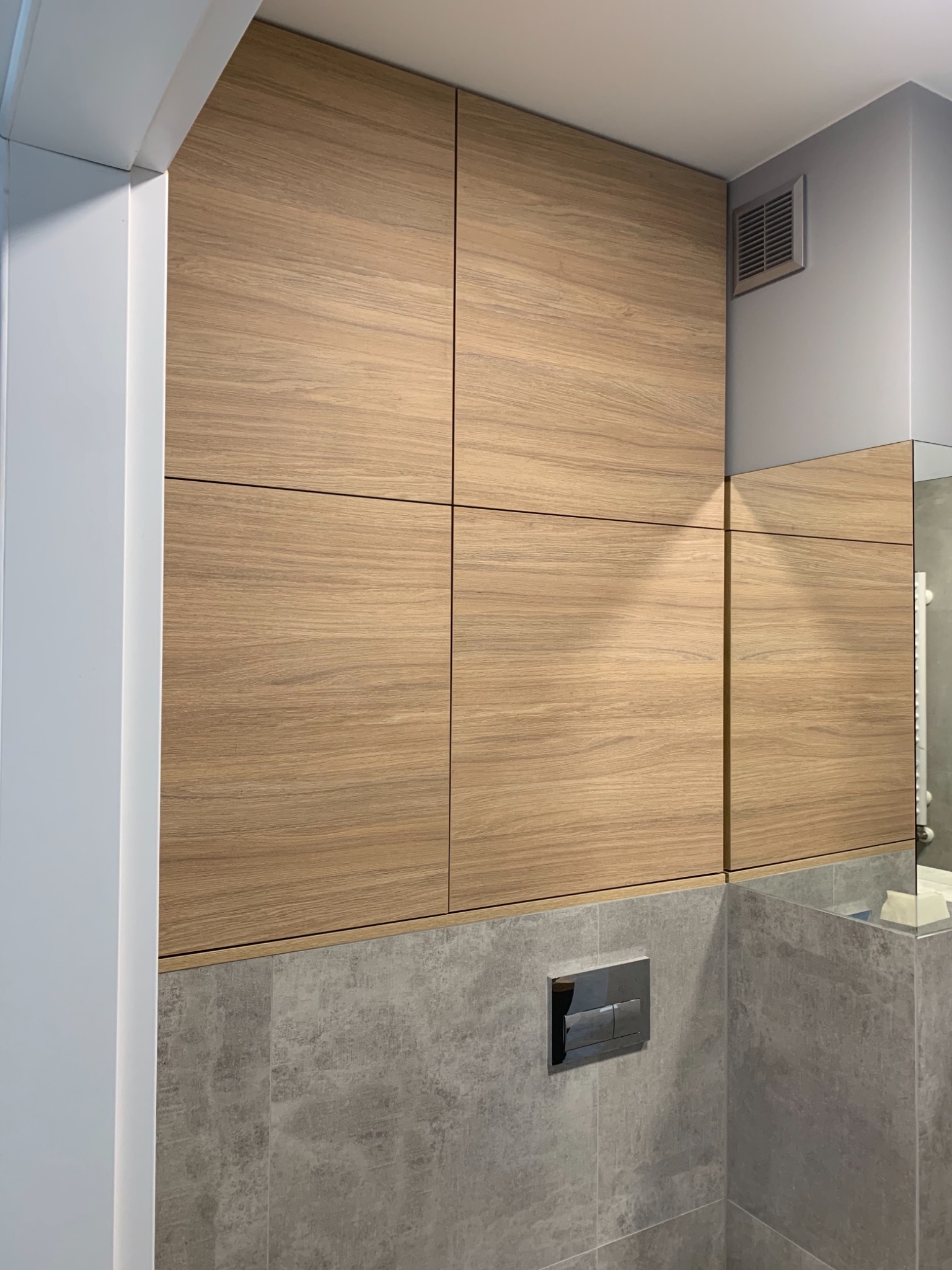 Drewniane szafki w zabudowie nad toaletą w łazience, z widocznym przyciskiem spłuczki i fragmentem lustra.