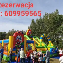 Kolorowy dmuchany plac zabaw z motywem klauna, otoczony grupą dzieci i dorosłych na tle zieleni i błękitnego nieba, z widocznym numerem telefonu do rezerwacji.