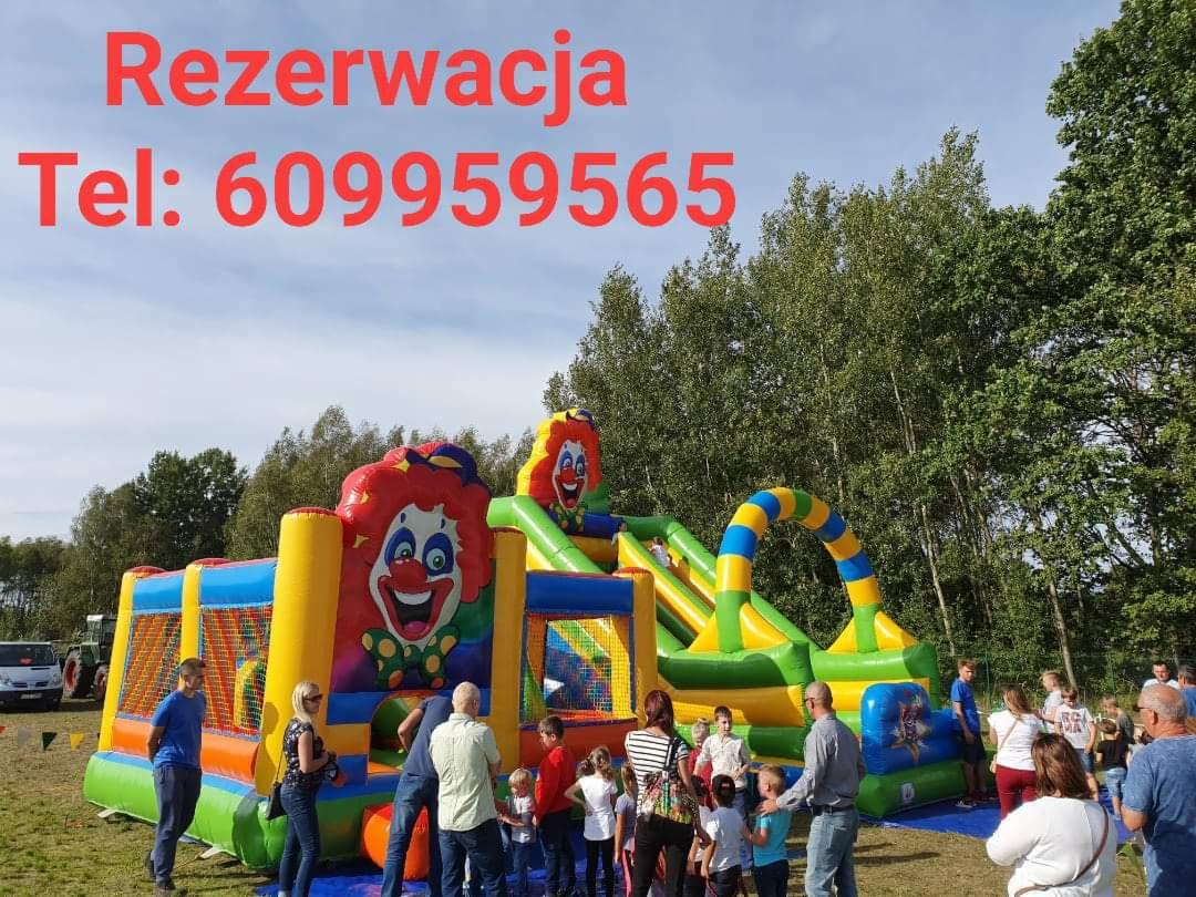 Kolorowy dmuchany plac zabaw z motywem klauna, otoczony grupą dzieci i dorosłych na tle zieleni i błękitnego nieba, z widocznym numerem telefonu do rezerwacji.