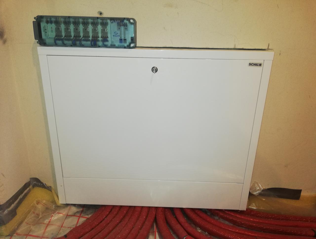 Rozdzielacz ogrzewania podłogowego marki Schilie z termostatami Salus, widoczne czerwone rury instalacji grzewczej na folii izolacyjnej.