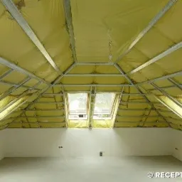 Poddasze w trakcie izolacji: widoczne metalowe profile, żółta wełna mineralna pomiędzy nimi oraz dwa okna dachowe w skosie dachu, ściany pomalowane na biało.